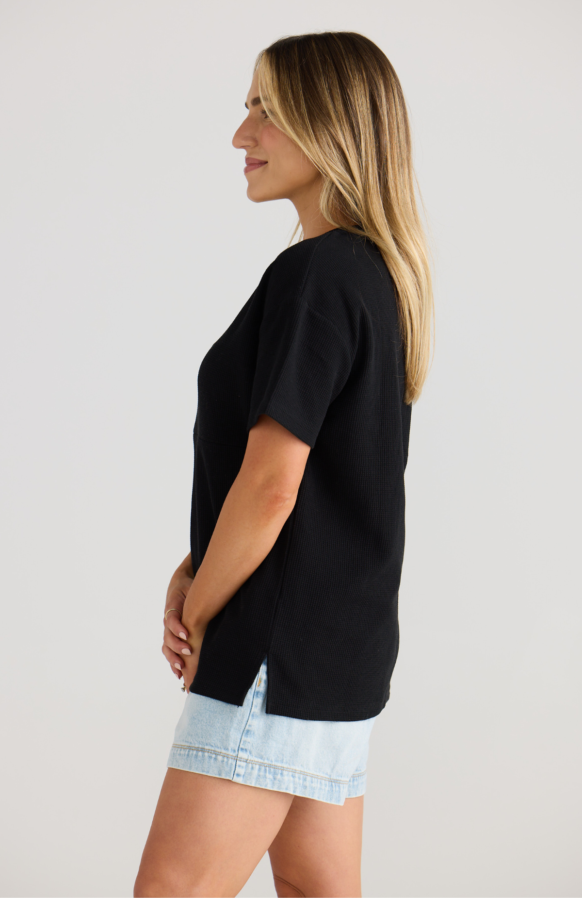 Woman wearing a black oversized breastfeeding-friendly t-shirt and light blue shorts on a plain background