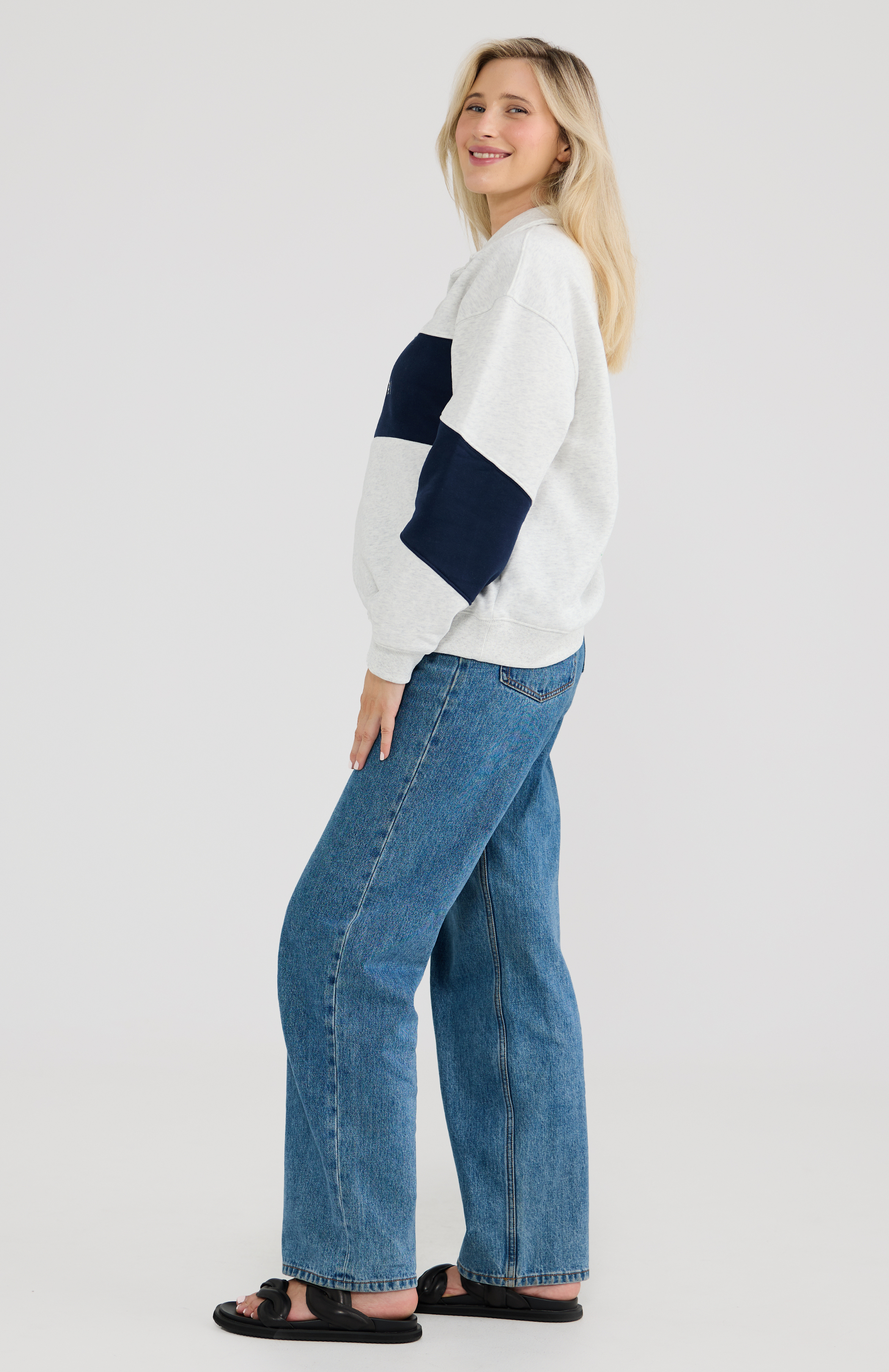 Woman wearing a grey and blue breastfeeding sweater with blue jeans on a light grey background