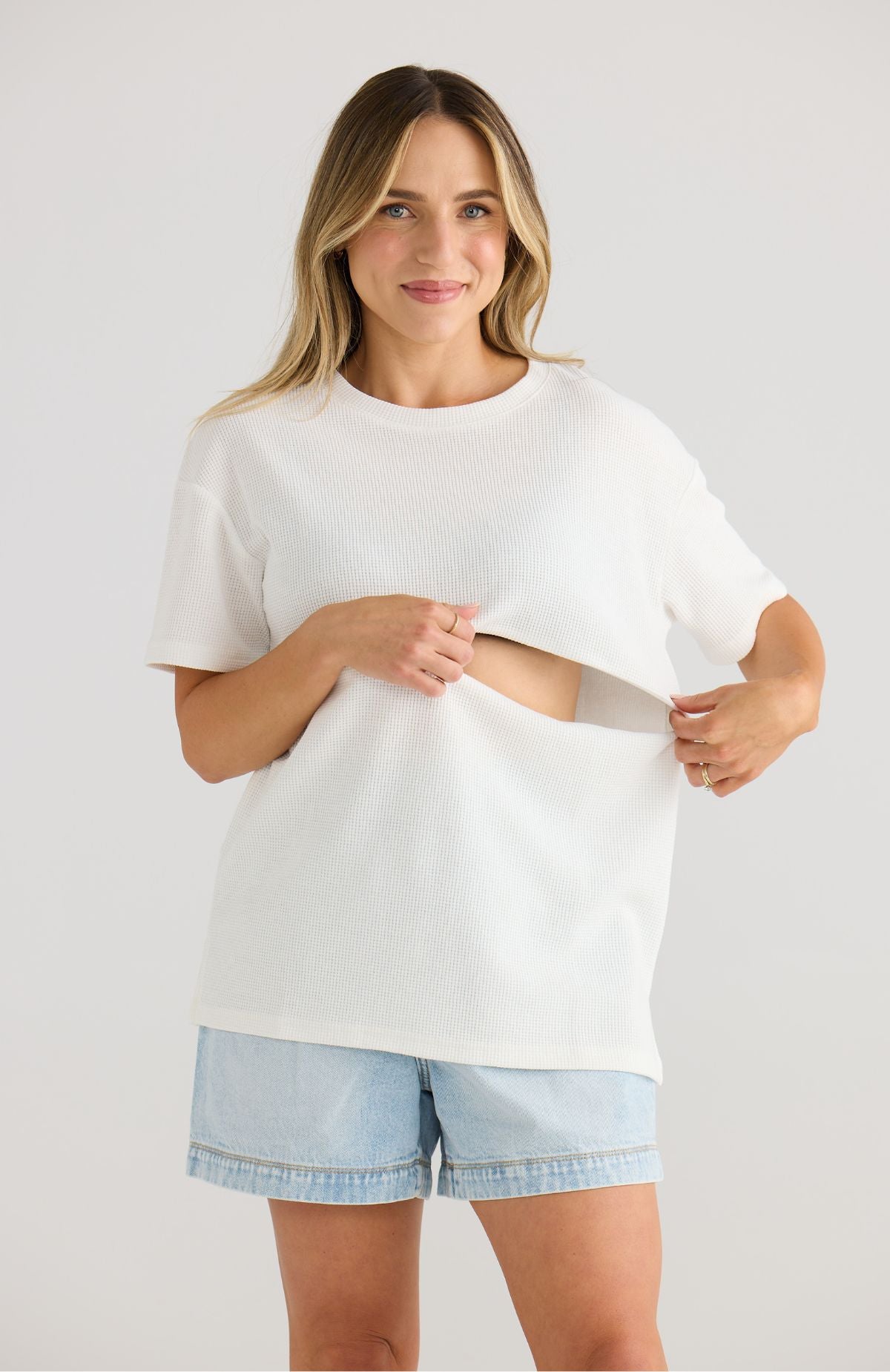 Woman wearing a white oversized breastfeeding t-shirt and light blue shorts on a plain background