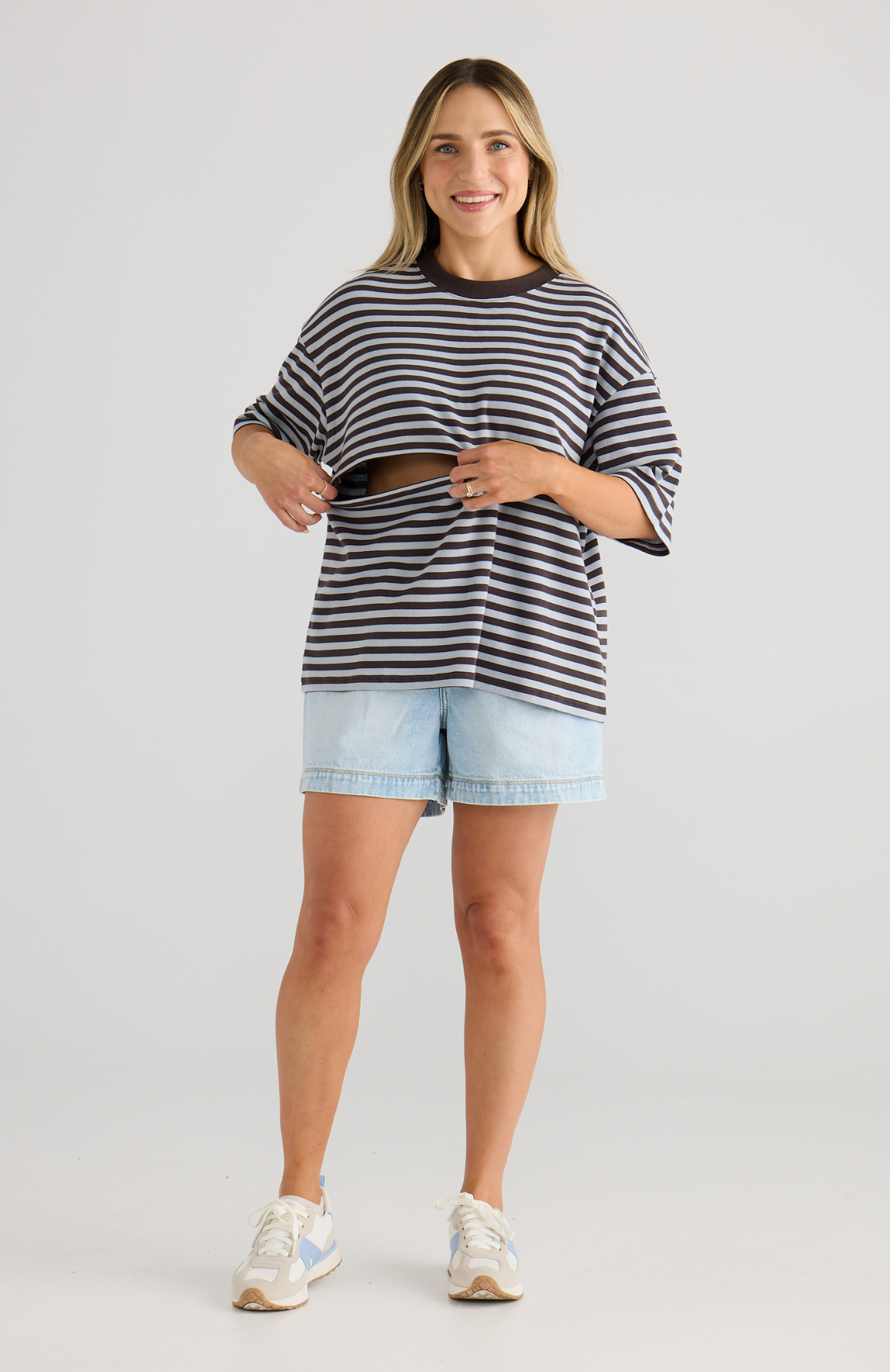 Woman wearing a chocolate and blue striped top and light blue shorts on a plain background