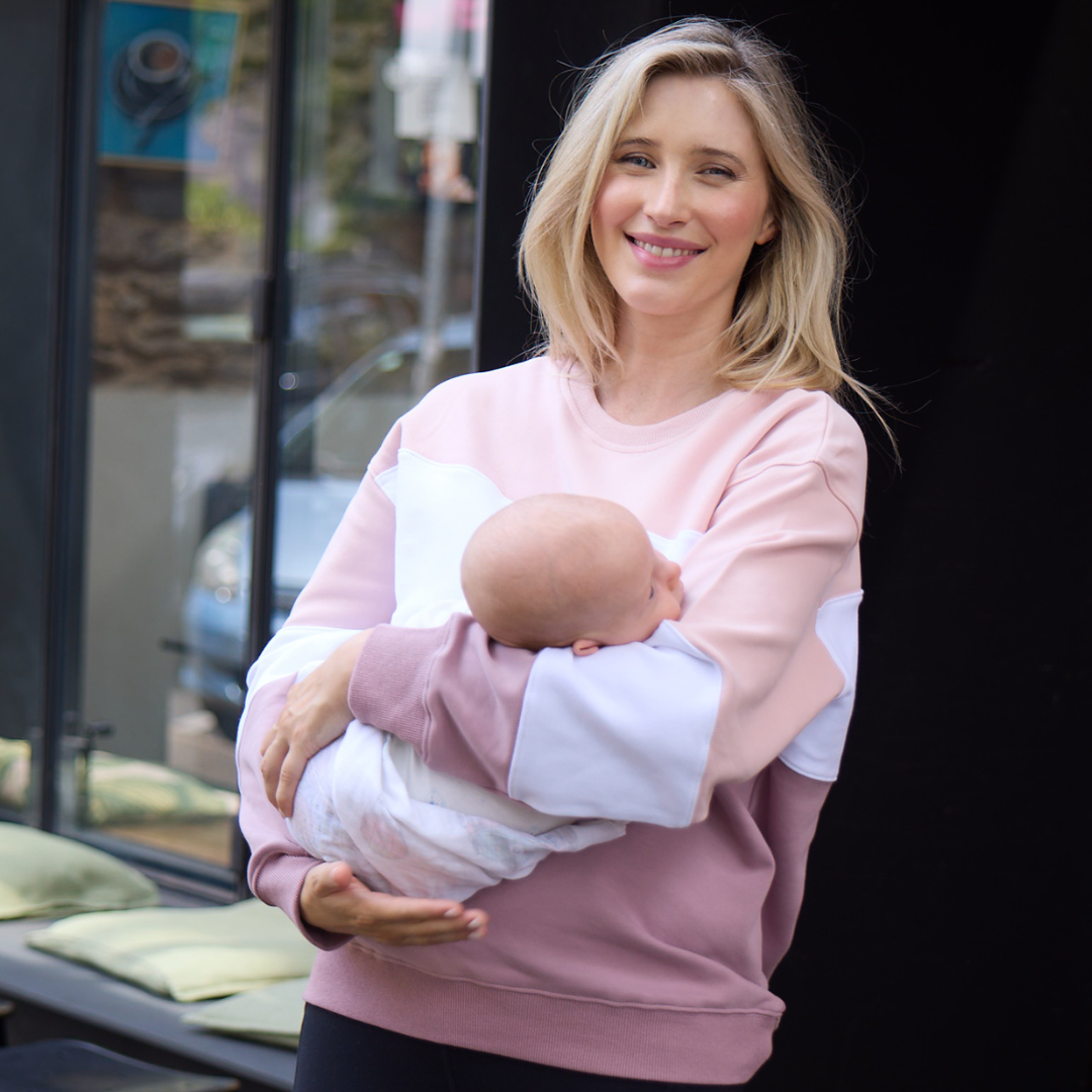 Woman holding a baby wrapped in a white blanket outdoors.