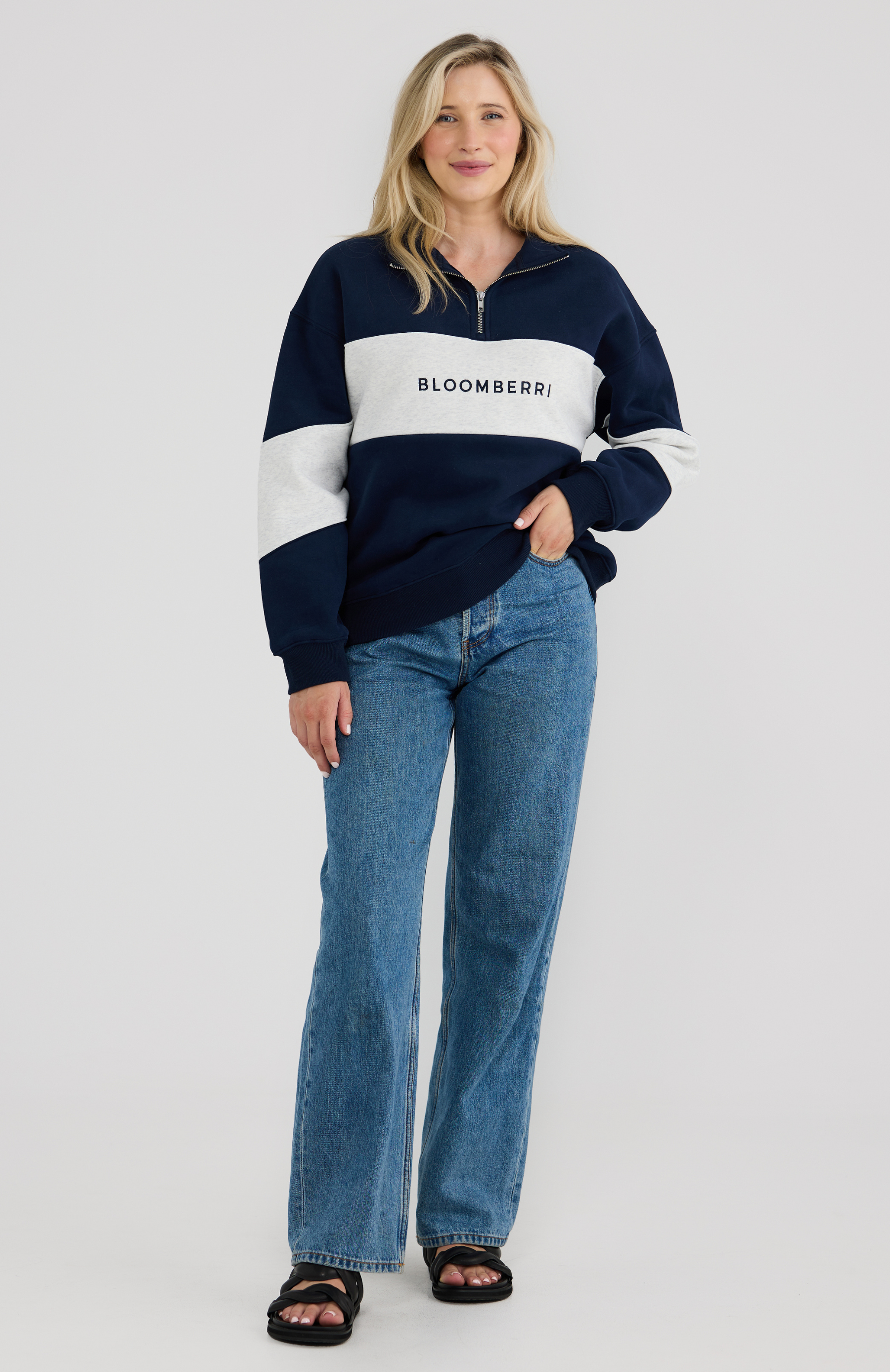 Woman wearing a navy and grey striped sweater with 'Bloomberri' logo and blue jeans on a light grey background