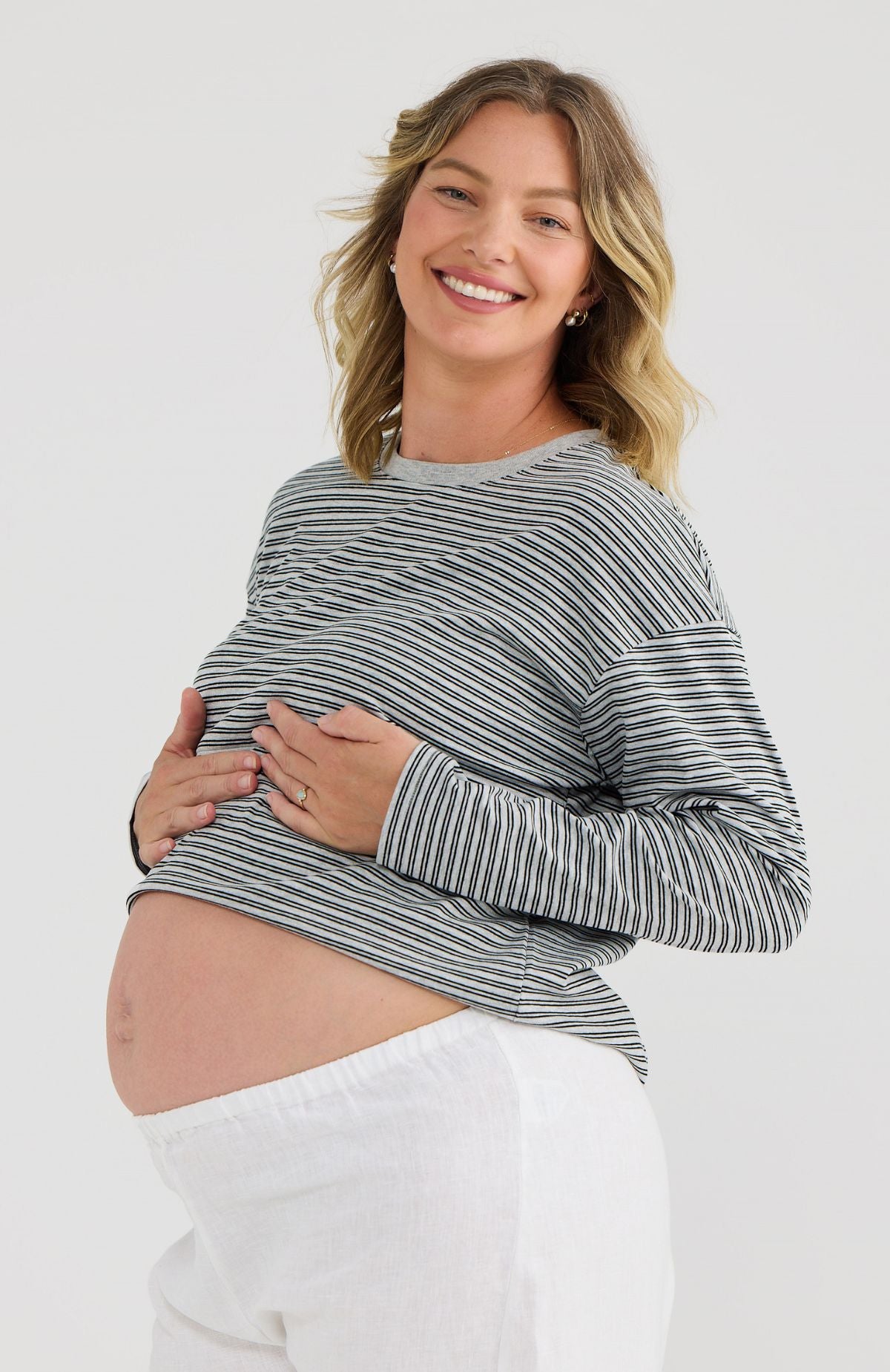 Pregnant woman wearing a striped long-sleeve top and white pants on a plain background