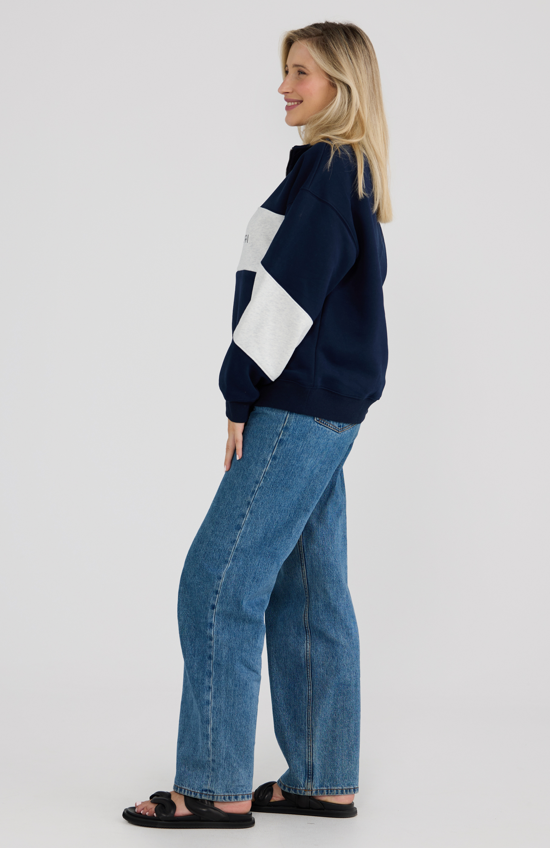 Woman wearing a navy sweater with a white sleeve panel and blue jeans on a light grey background