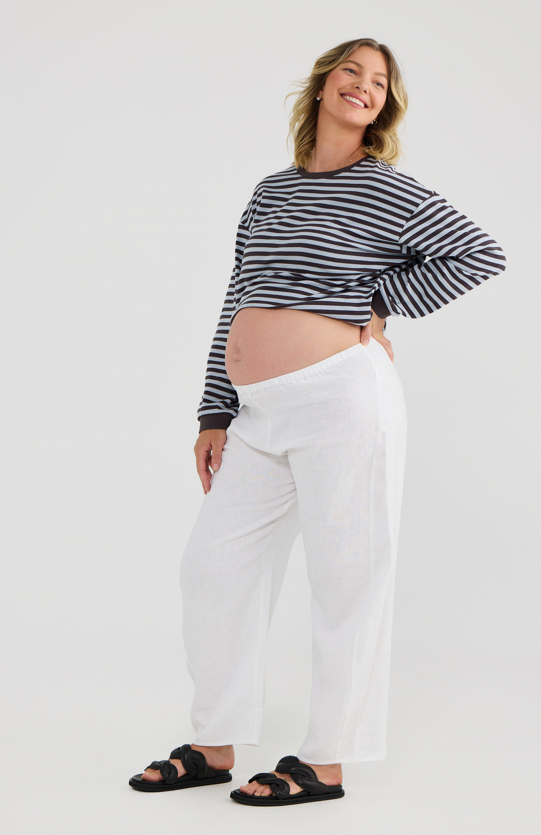 Pregnant woman wearing a striped long sleeve top and white pants on a plain background